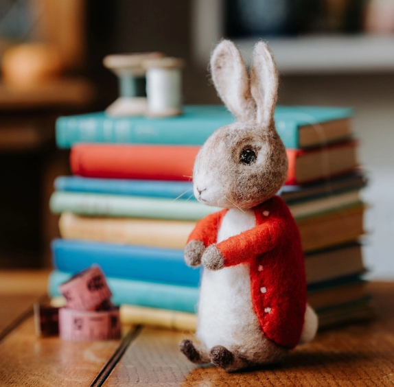 Close up of the felted Bertie Bunny in a red jacket standing in front of a stack of books. 