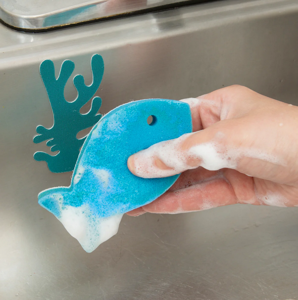 The blue fish shaped scrubber filled with suds with the blue coral shaped holder attached to the sink in the background. 