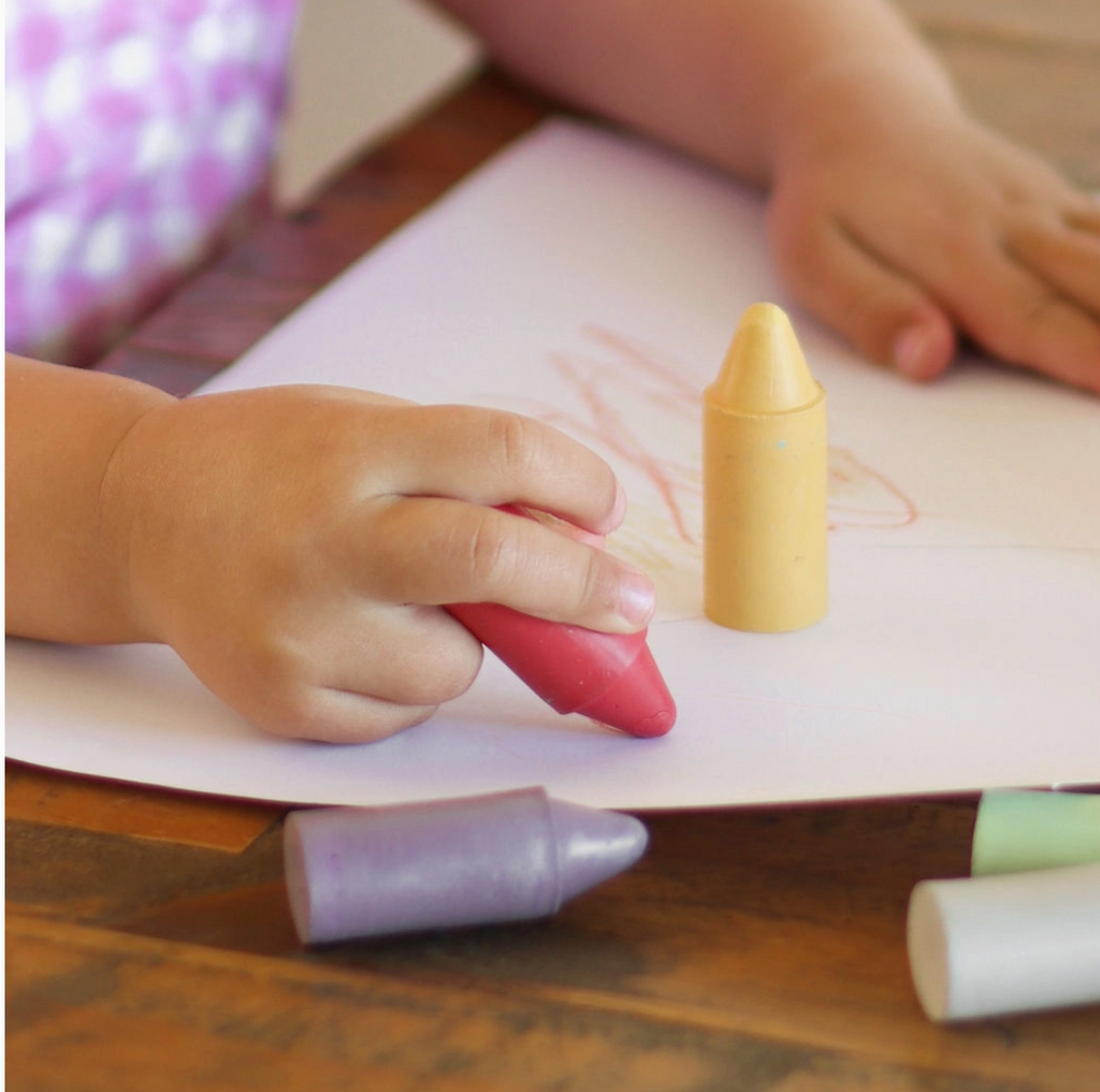 The hands of a child drawing with jumbo crayons on a piece of paper at a table.
