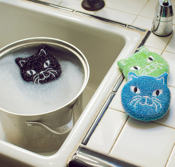 The black kitty scrubber floating in a pot of soapy water in the sink with the blue and green kitty scrubbers on the counter beside the sink. 