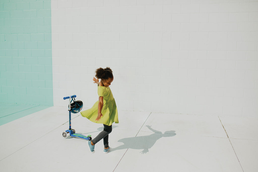 Happy young Black girl in a yellow dress, grey leggings, and sneakers spinning while looking at her shadow next to a Crystal Blue LED Mini Scooter with a helmet on the handlebars.