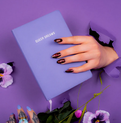 A hand coming through a dark purple wall holding the Queer Dreams journal. 
