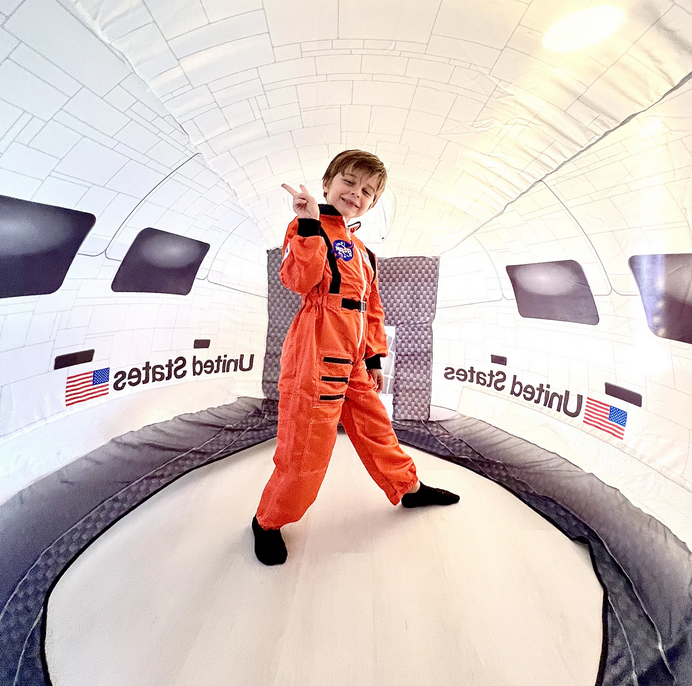 A young child dressed in an orange NASA flight suit standing inside the Space Shuttle AirFort. 