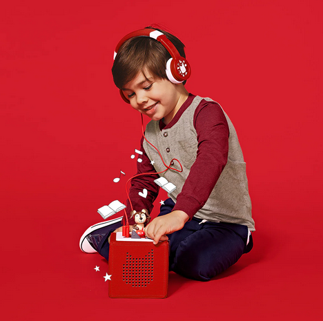 A young child sitting with the red Toniebox and listening to Playtime Puppy wearing the red headphones.  