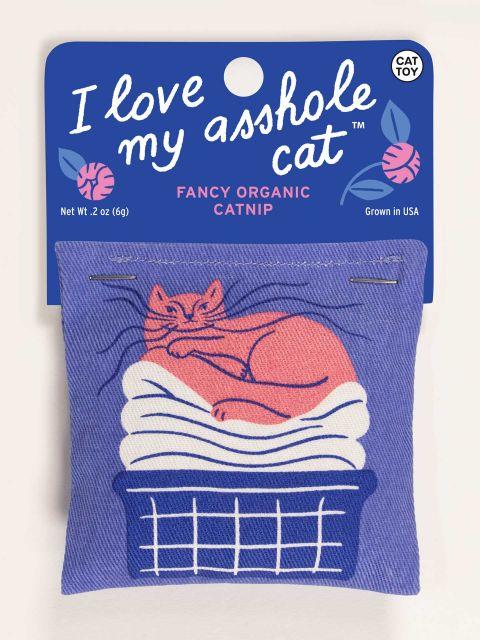 Front of denim blue catnip toy with a pink cat laying on top of clean laundry in a blue and white basket. the toy is attached to a cardboard hang tag that is blue with pink flowers and the words " I love my asshole cat" in white cursive letters. 