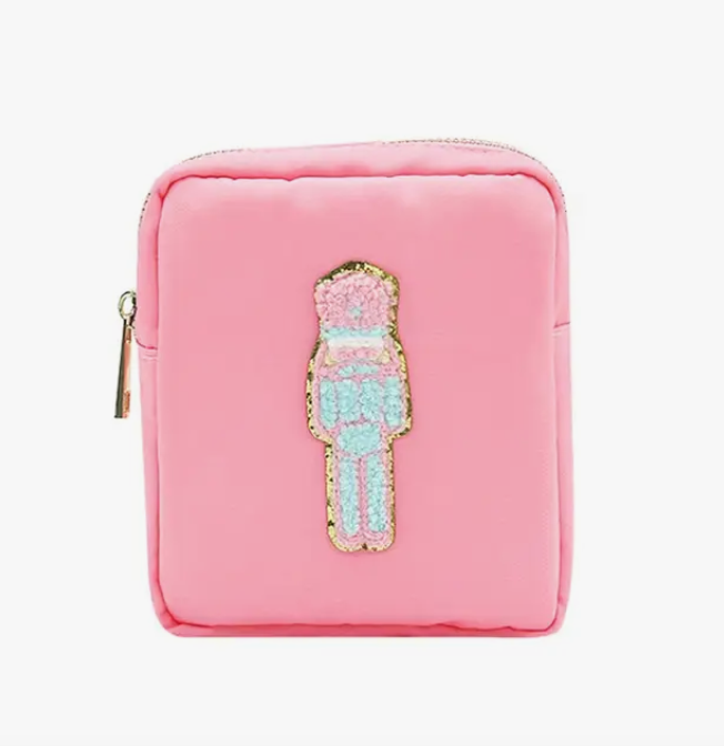 A cute pastel nutcracker embroidered in fluffy chenille and outlined in gold on a bag that is soft pink with a bright gold zipper. 