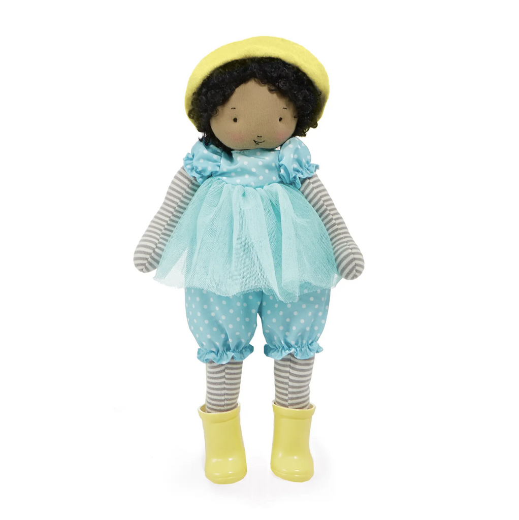 Plush brown skinned doll with curly brown hair. Phoebe is wearing a blue and white polkadot jumpsuit with a blue tulle skirt, a yellow felt hat, and yellow rain boots.