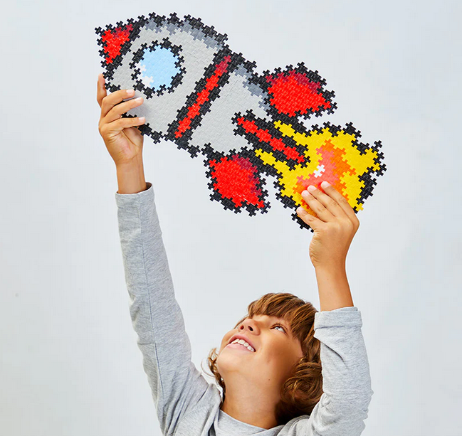 A child playing with a completed Plus Plus Puzzle By Number Rocket. The rocket is held high above his head as if its flying through space.  The completed puzzle is white and red with yellow and orange flames projecting it through the universe. 