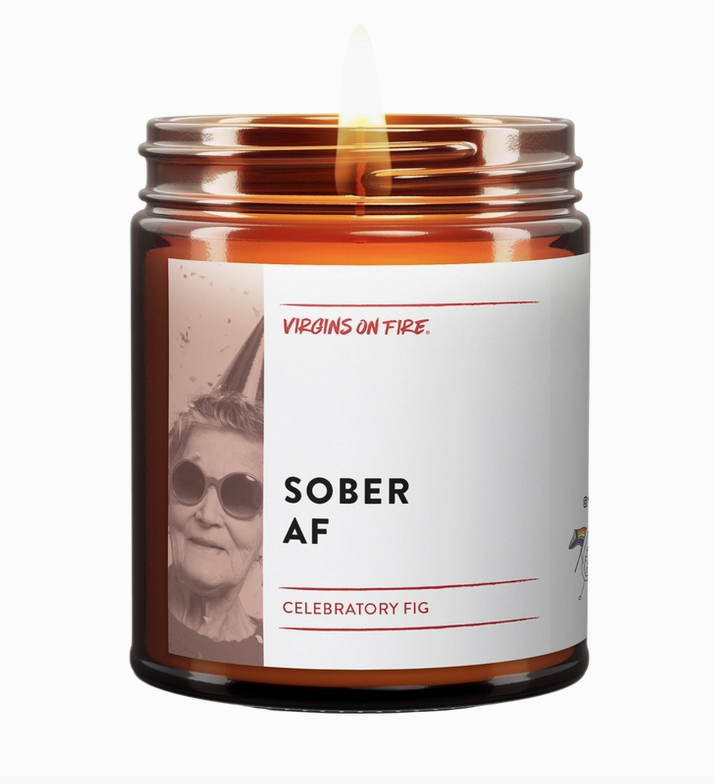 Soy wax candle in a brown glass jar with flame. It has a white wrap around label with black lettering that reads "Sober AF" with a black and white photo of an older woamn wearing sunglasses. 