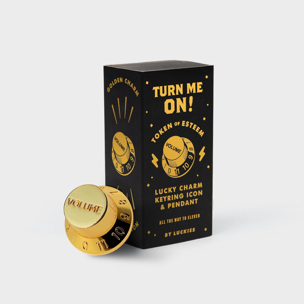 Black box with gold foil lettering and graphics that read "Turn Me On" "Token of Esteem" and "Lucky Charm Keyring Icon & Pendant" The gold metal token shaped like a knob is leaning against the box. 