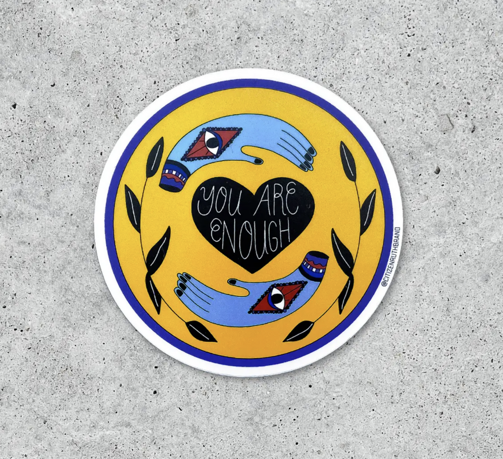 Round yellow sticker with a black heart that reads You Are Enough in white letters in the center surrounded by two blue hands with eyes in the palm and leaves.