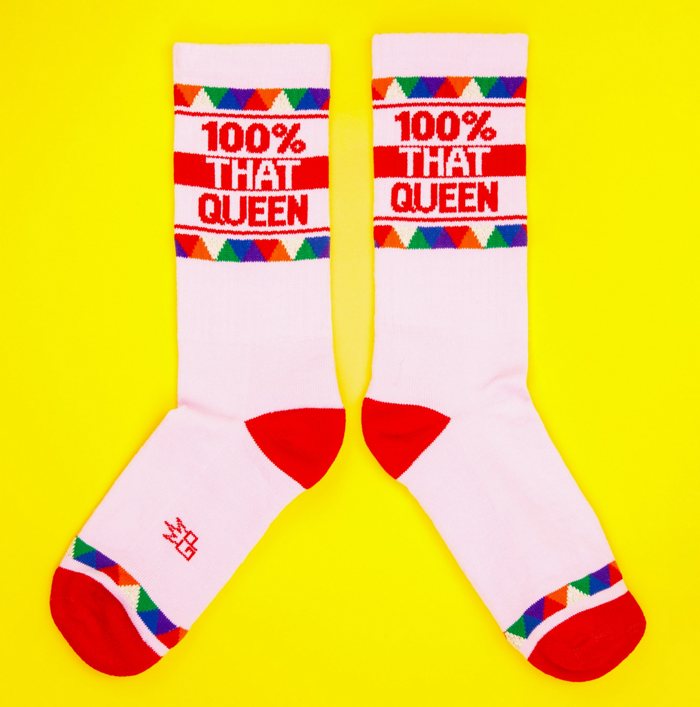 A pair of light pink tube socks with red and multi colored stripes that read "100% That Queen" in red letters. 