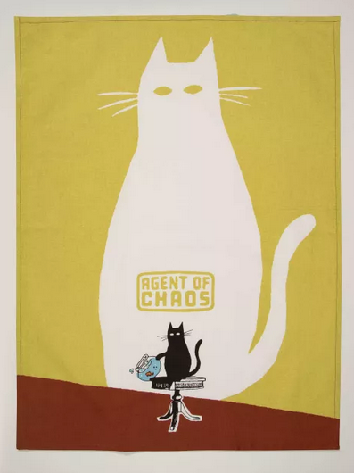A greenish yellow dishtowel laid flat with a sihouette of a large white cat in the background and a black cat on a table knocking off a fishbowl. The words "Agent of Chaos" are printed above the black cat.  