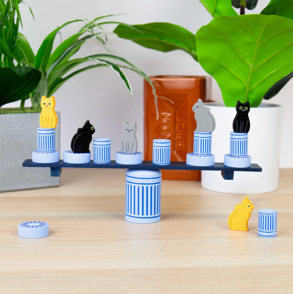 Balancing toy with cat figurines and trash cans on a wooden surface with plants in the background
