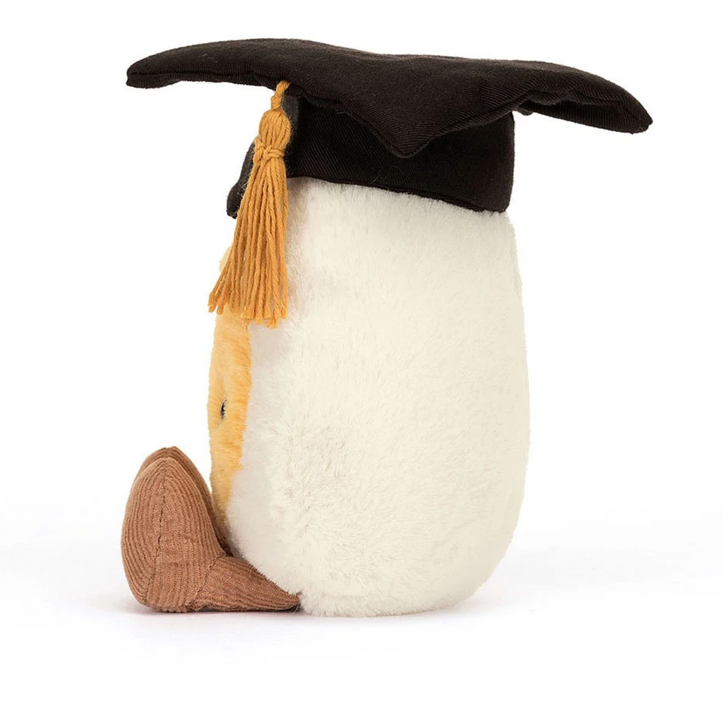 View from left side of plush boiled egg wearing a graduation cap with gold tassel.