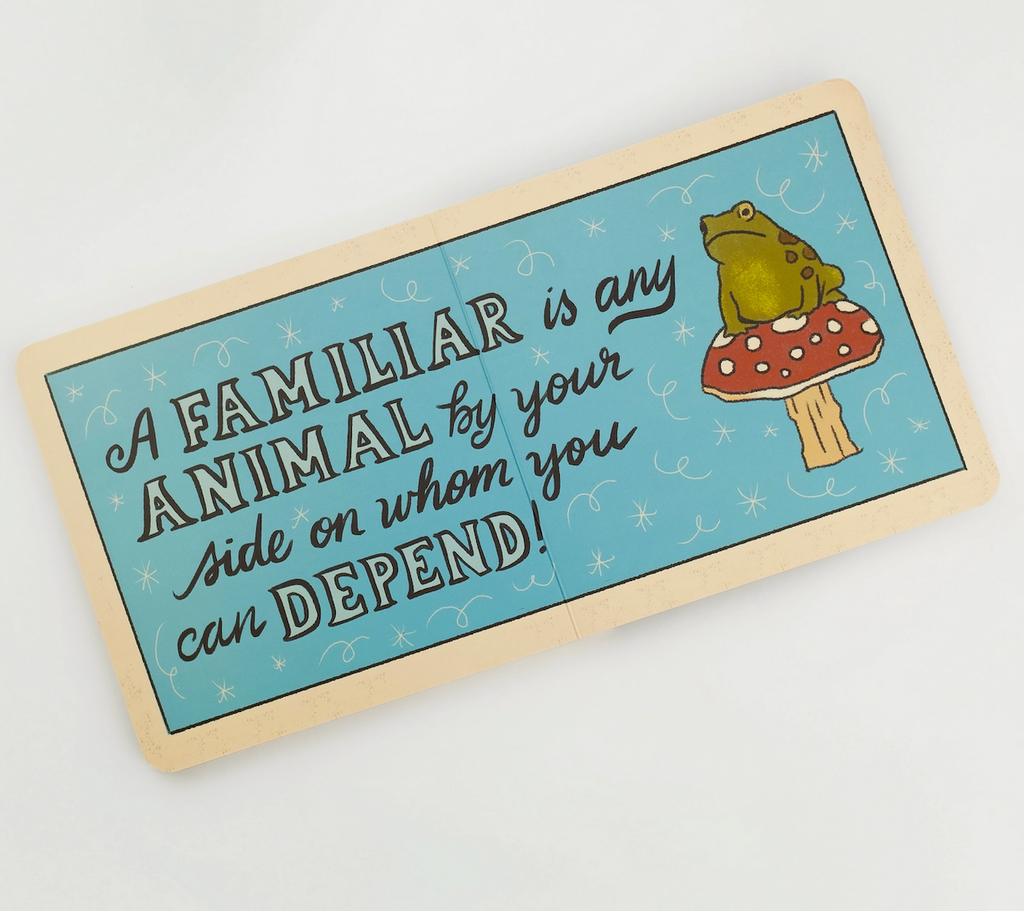 Baby's Book of Familiars board book opened to an interior page with an illustrated frog sitting on top of a mushroom that reads "A familiar is any animal by your side on whom you can depend!"