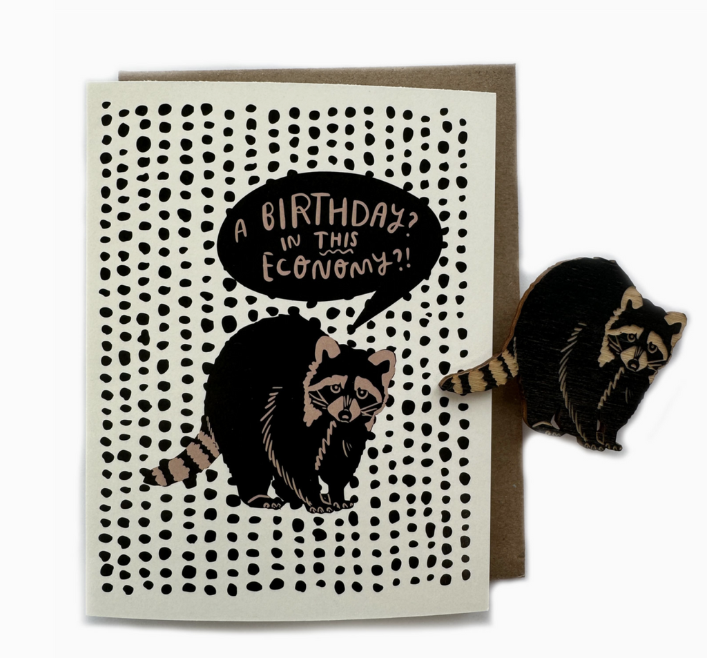 Greeting card with black dots repoeating pattern and a raccoon with a word bubble that reads "A Birthday in this Economy?" A magnet of the raccoon is off to the side.