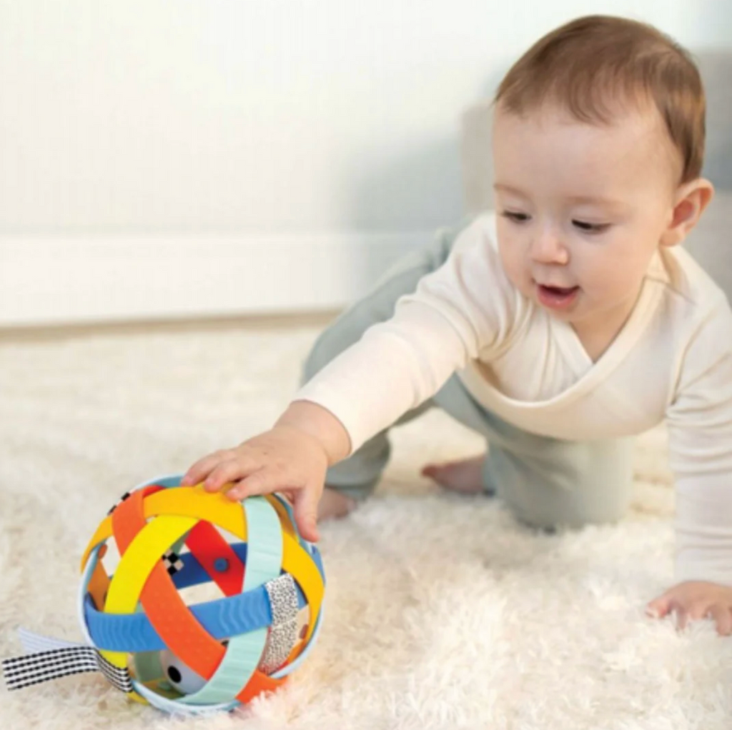 A baby  crawling and reaching for the Blissful Ball which is a colorful ball with soft, colorful, textured bands that are open to show a smaller ball inside. 