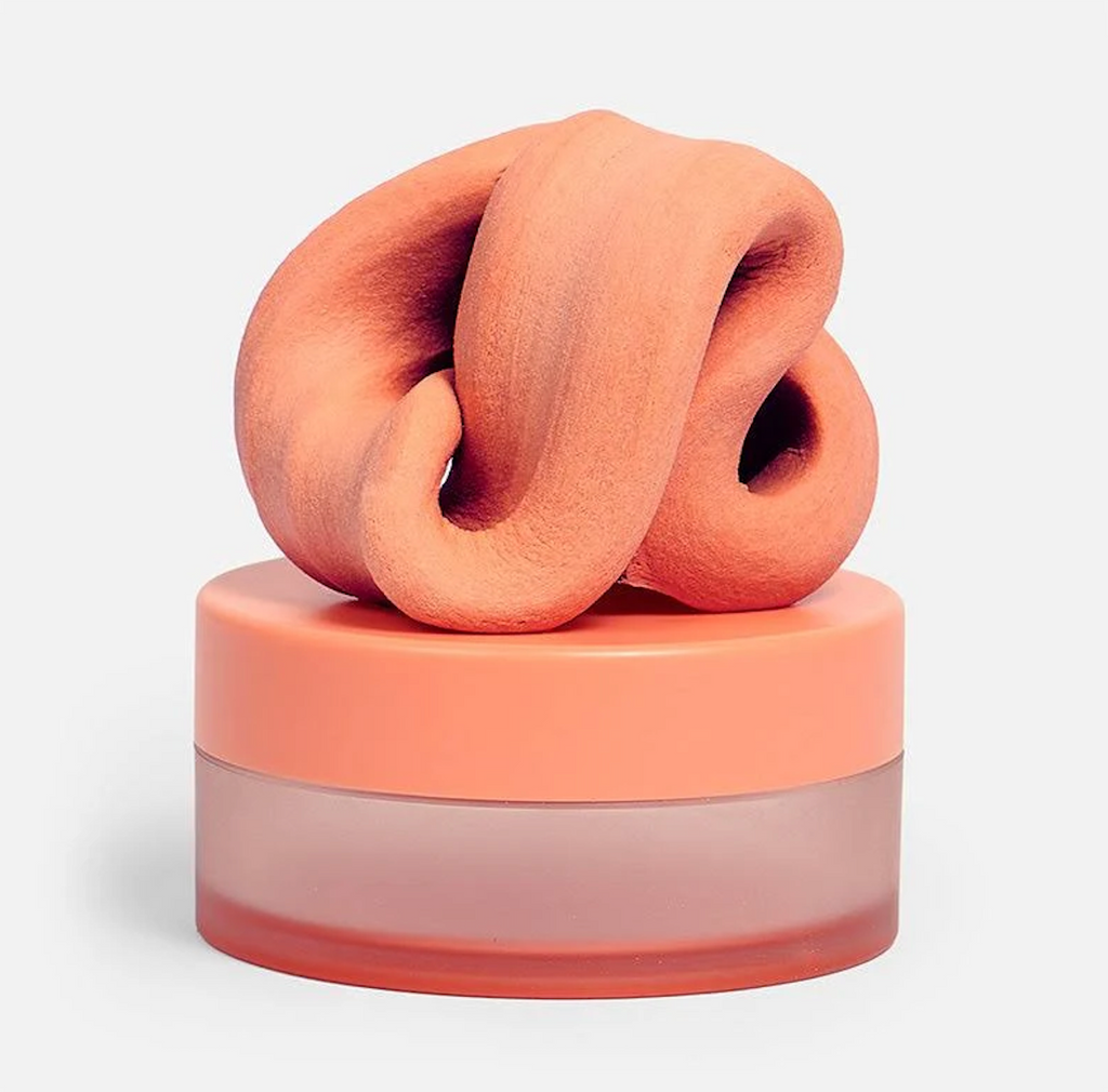 A pile of the Blur Focus Putty from Speks in the color Clementine a soft orange sitting on top of the matching container it is packaged in. 