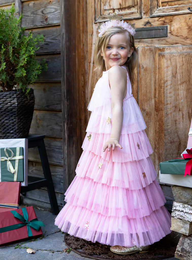 A child wearing the Blush Christmas Tree Dress with pink tulle in three shades with glitter stars, ornaments and candy canes throughout. She is also wearing the matching headband with three gold, glitter stars atop a pink tulle wrapped headband. 
