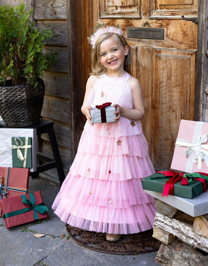 A young child holding a beautifully wrapped present while wearing the Blush Christmas Tree Dress and matching headband that has three gold glittery stars attached to a headband wrapped with pink tulle. 