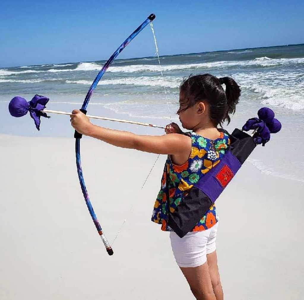A kid on a beach with the Two Bros. Bows aiming her arrow at a target in the distance. 