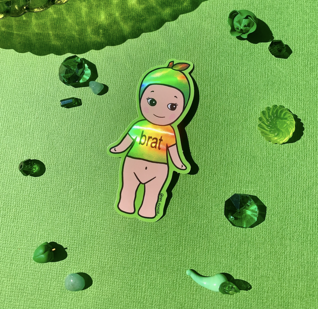 Holographic sticker of a kewpie doll with a green t-shirt and hat that reads BRAT