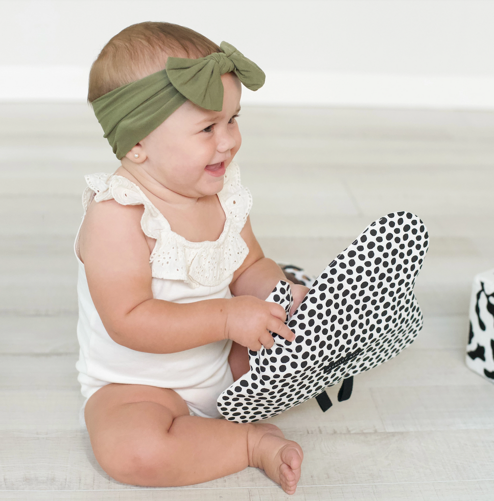 A baby sitting up holding the Butterfly Crinkle Toy showing the back side which is white with black polka dots. 