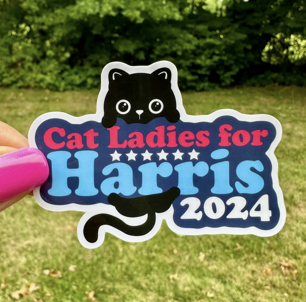 A shaped vinyl sticker with a cute black cat hanging on lettering that reads "Cat Ladies for Harris 2024"