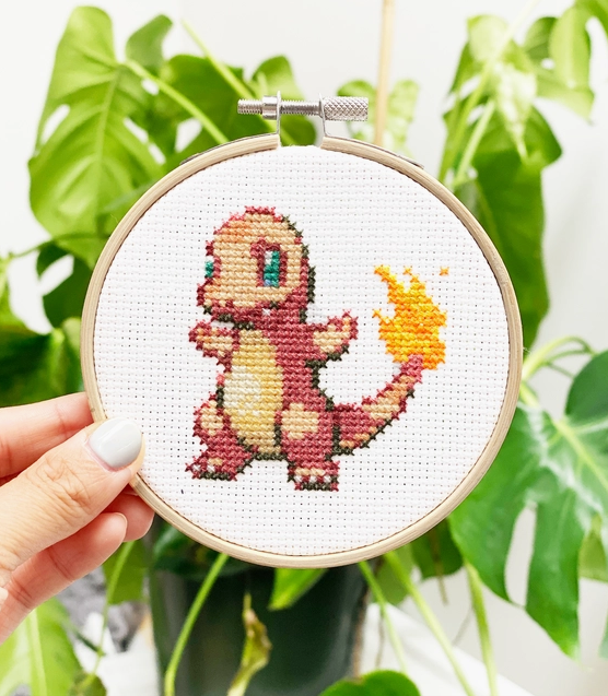 A completed Charmander cross stitch framed in the wooden hoop being held in a hand for size scale. 