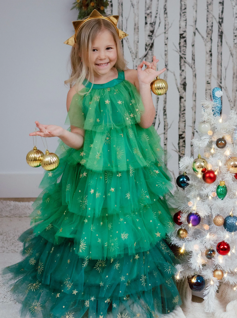A young child wearing the Christmas Tree Dress and headband while standing beside a white Christmas tree and holding gold ornaments. 