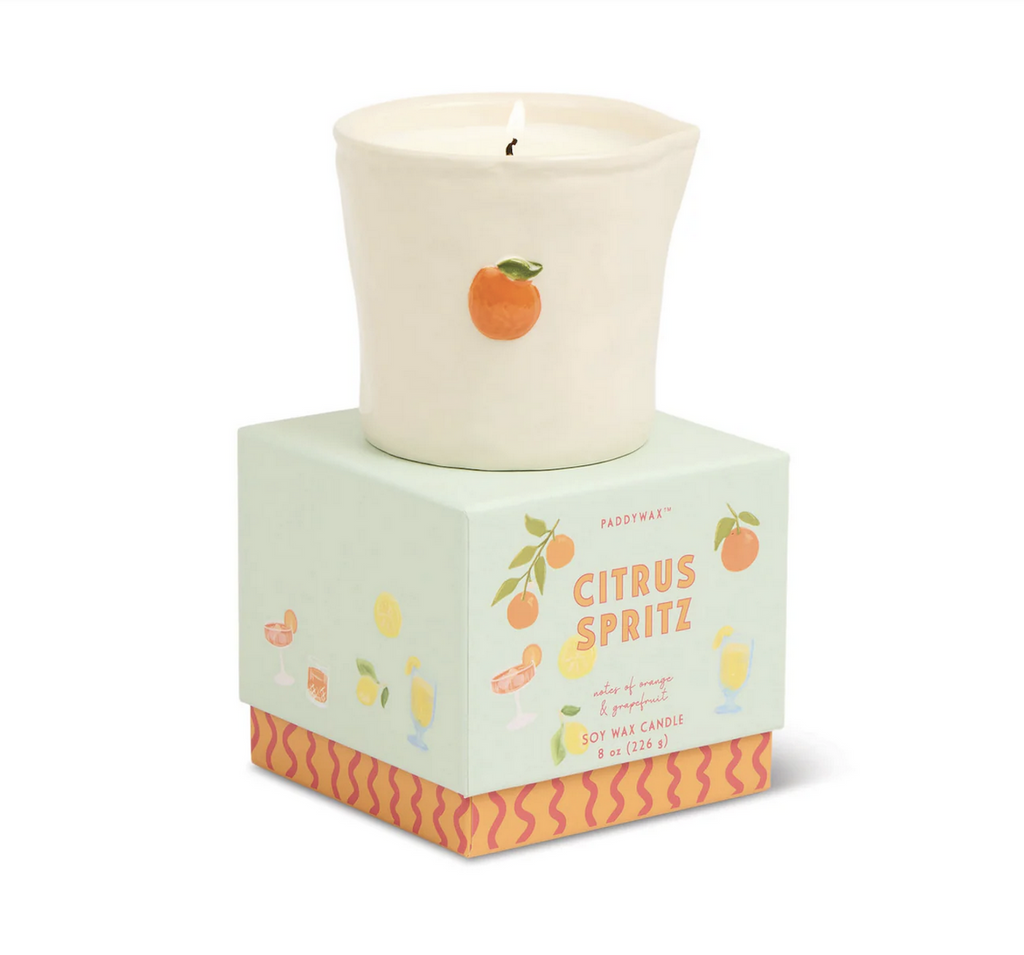 Citrus Spritz scented candle from Paddywax in a reuseable, food safe vessel with a hand painted orange on the front. The candle is lit and sitting on top of the beautifully decorated gift box that accompanies it. 