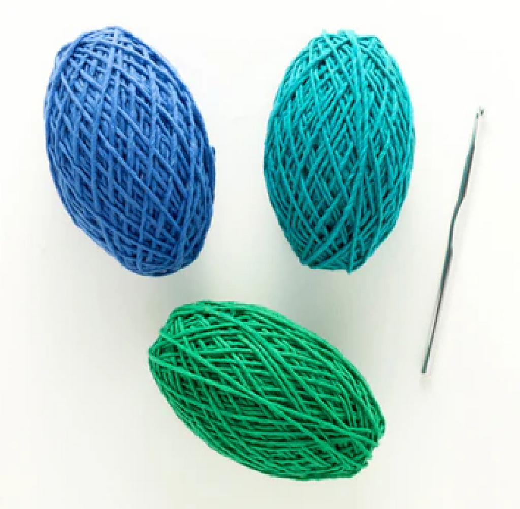 Three balls of yarn in blue, green, and teal with the crochet hook that comes inside the crochet kit. 