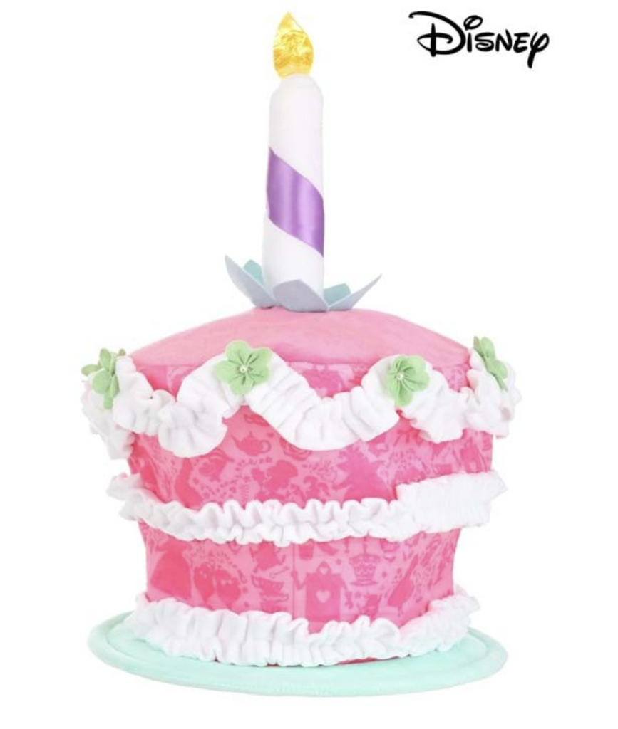 Plush pink cake hat with a single candle on top. There are ribbons of white icing separating the layers and green flowers decorating the top. 