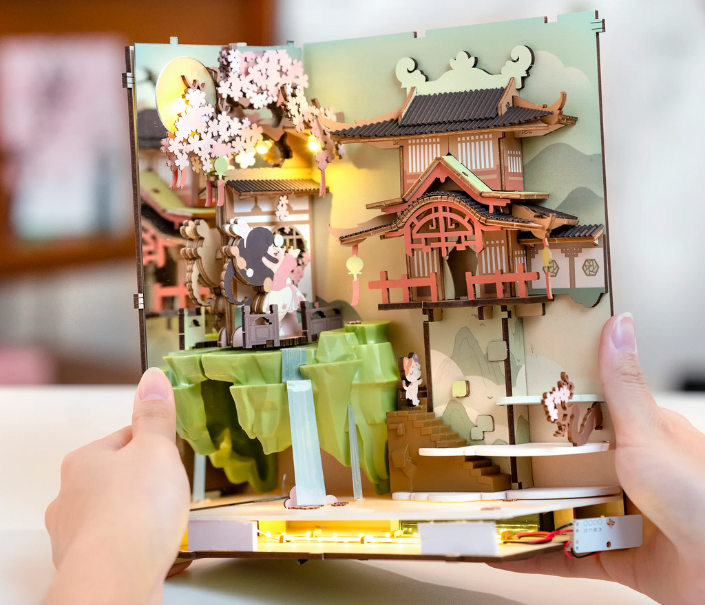 Interior scene shows book nook kit partially completed, with a girl frolicking in the falling cherry blossoms, a waterfall, cat on stairs going up to another pagoda. LED lights can be seen underneath.