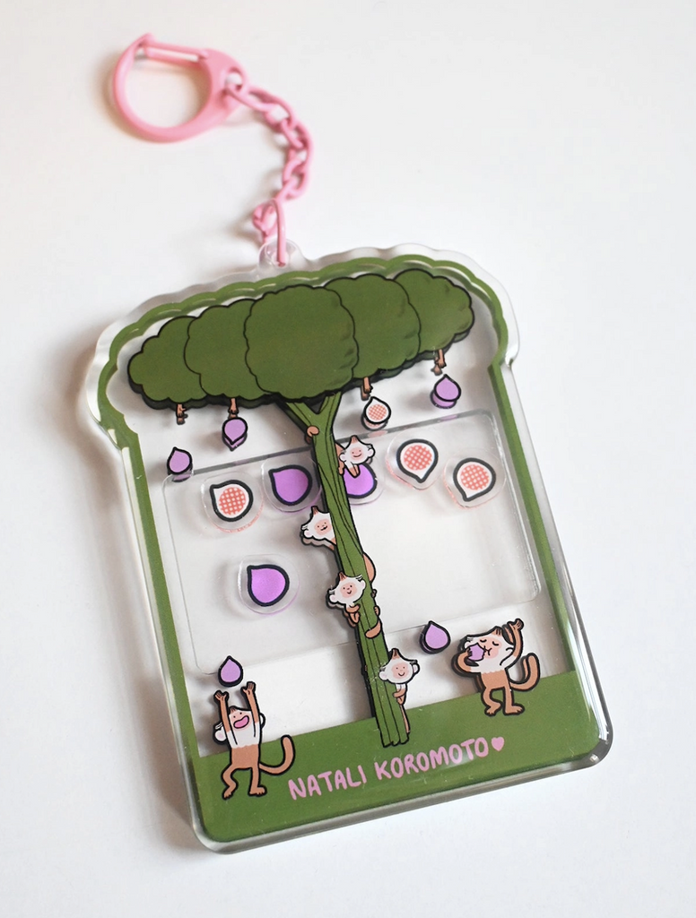 A clear acrylic keychain that has a green tree with figs falling from it and capuchin monkeys chasing after the figs. 