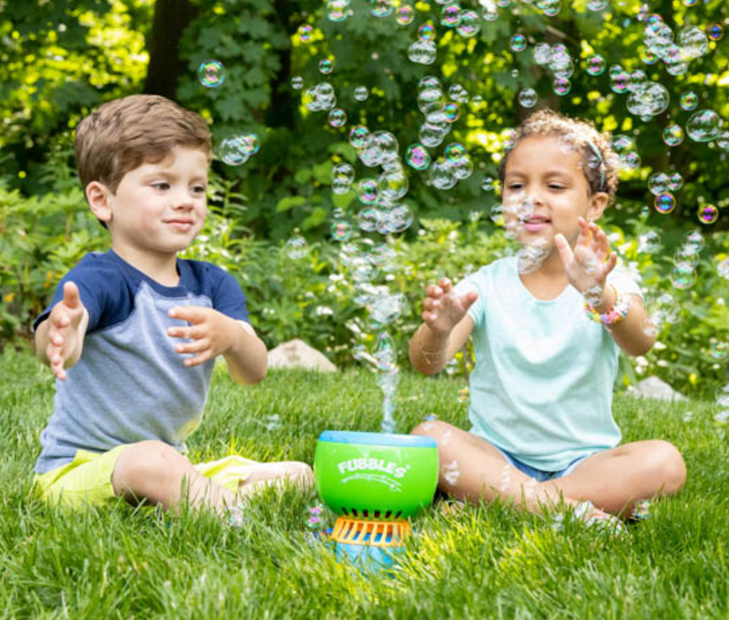 To children sitting in the grass around the Fun Finiti bubble machine filling the world with bubbles. 