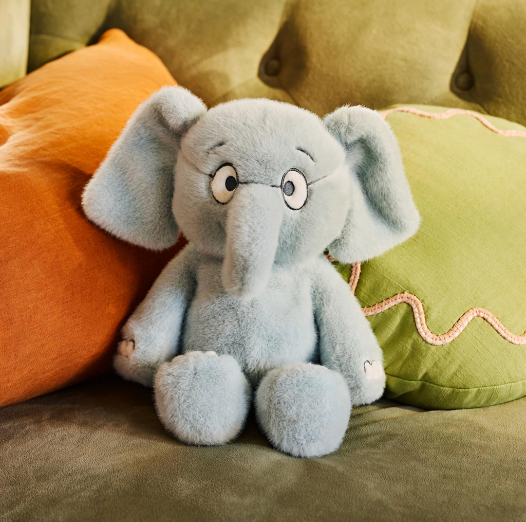 Gerald the Elephant plush a character from Mo Willems' books snuggled on a couch with orange and green pillows. 