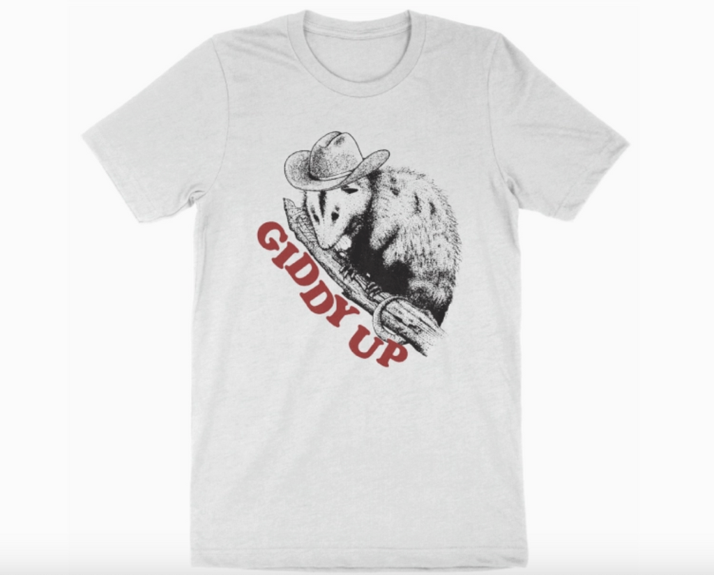 Illustrated sketch of a possum perched on a branch wearing a cowboy hat that reads "Giddy Up" in red letters, printed on a white t-shirt. 