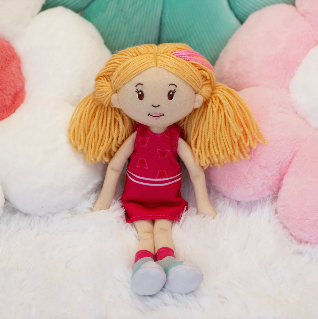 Groovy Girls doll Rose, with blonde yarn hair that has a pink streak sitting on a blanket surrounded by flower shaped pillows. 