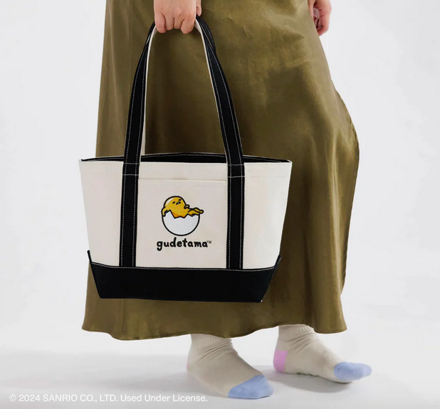 The Small Canvas Tote being held at the sideof an adult, showing the beige bag with navy handles and piping and Gudetama lazing in a broken egg. 