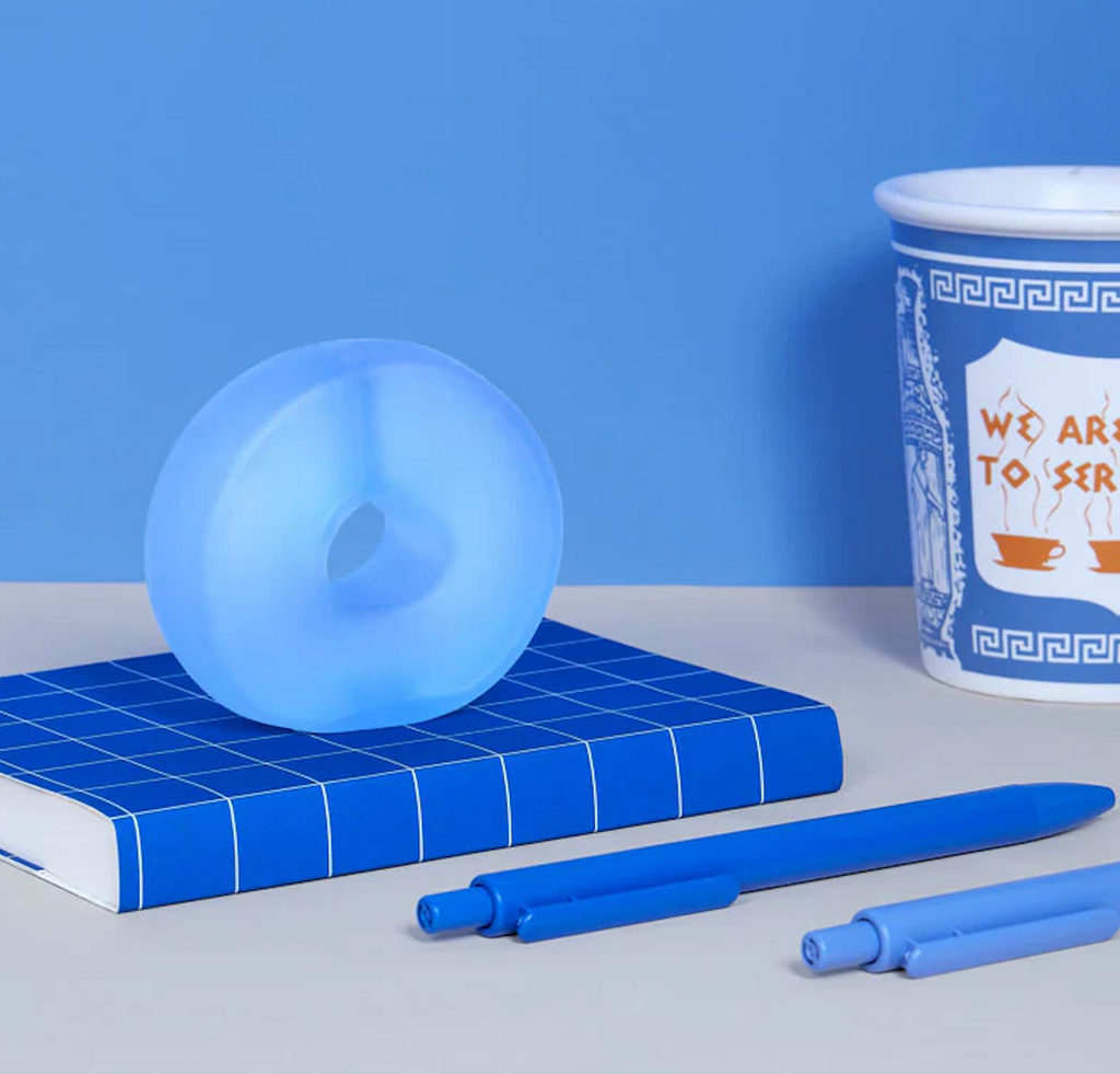 The Gump Loop in the color Dew, a squishy, stress toy sitting on a blue notebook on a desk beside some pens and a coffee cup. 