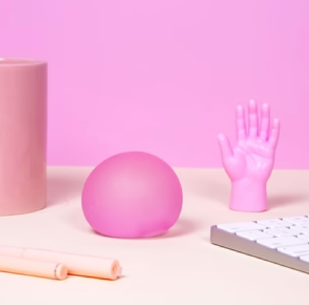 The Gump stress ball from Speks sitting on a desk by a keyboard and a pink hand in the color Moon Jelly. 
