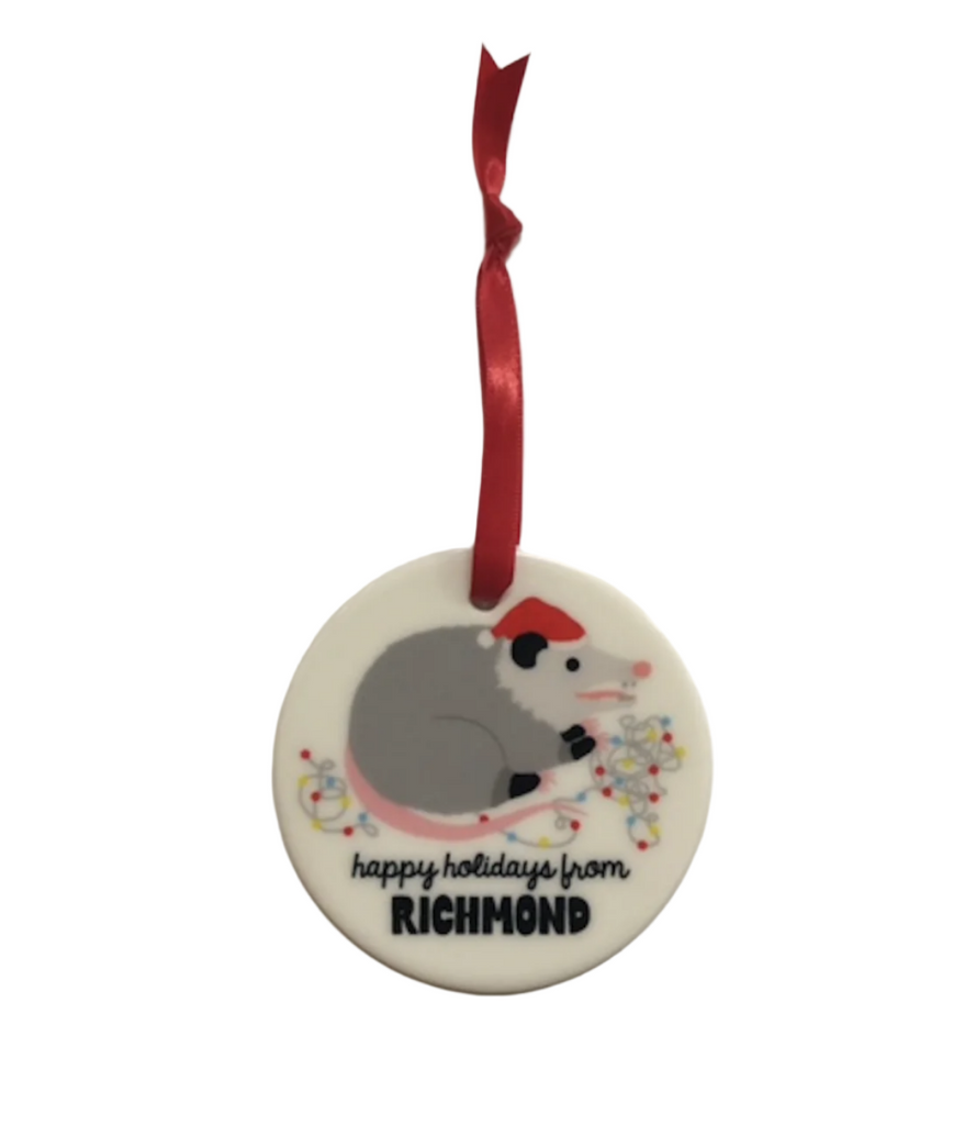 Ceramic disc ornament with illustration of an opossum wearing a Santa hat chewing a light string. 