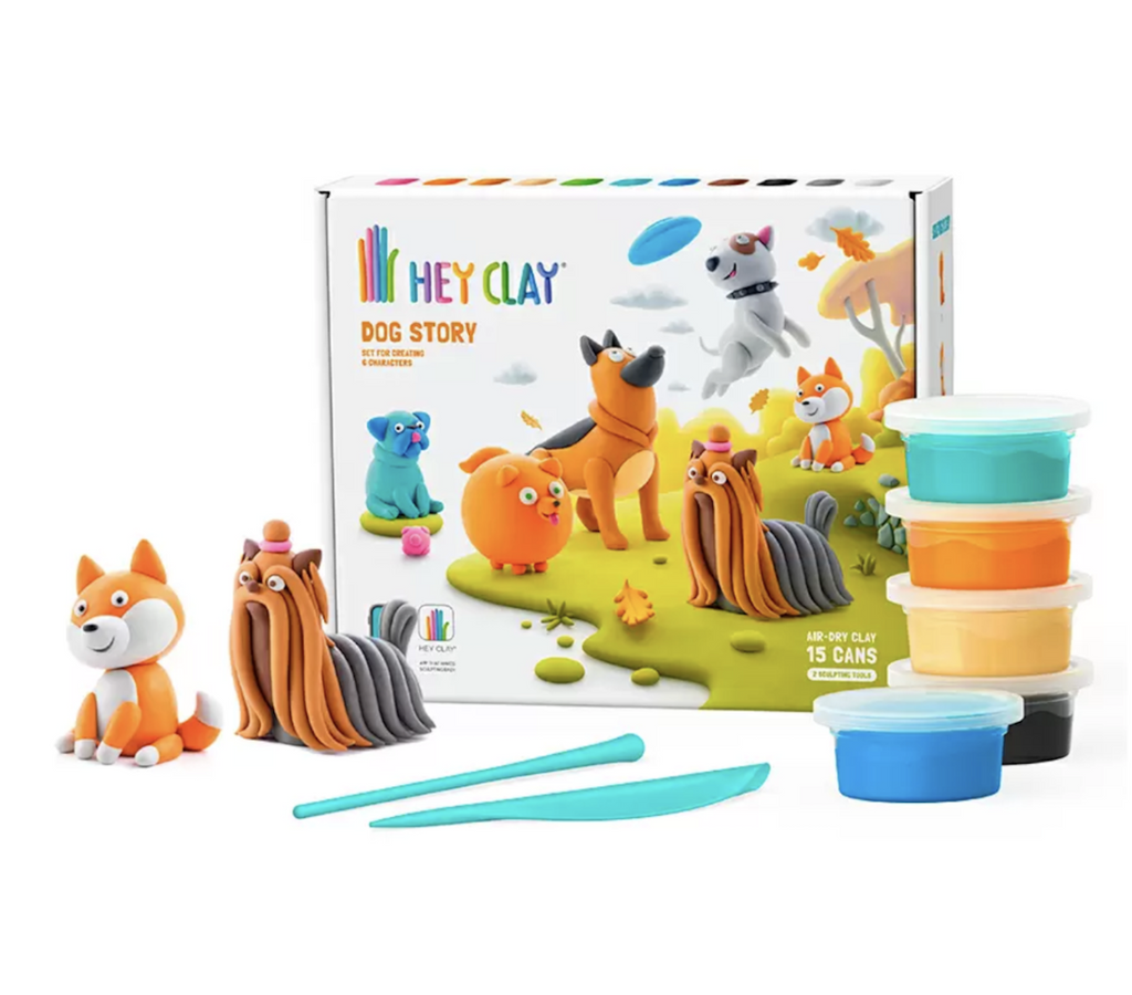Two dogs made from clay using the tools and clay from the Hey Clay Dog Story kit. There are sculpting tools and cans of colored air dry clay in front of the box, which has a picture of several dogs in an outdoor scene on the front. 