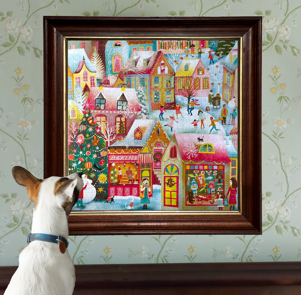 The completed Holiday Village 1K piece puzzle put together and framed with a furry friend admiring it on a wall. 