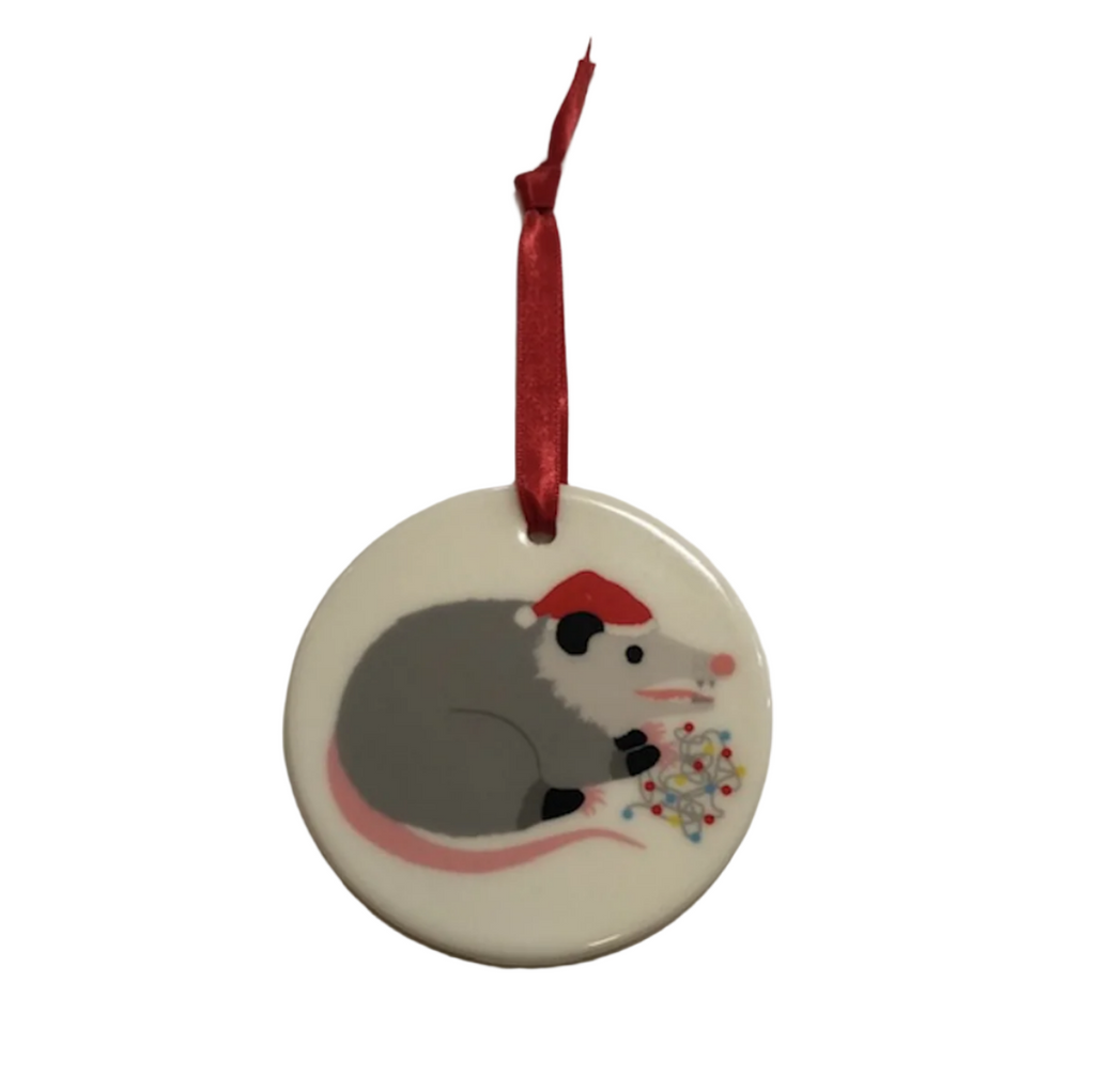 Ceramic, round ornament with red ribbon for hanging and an illustration of an opossum wearing a Santa hat and chewing on string lights. 