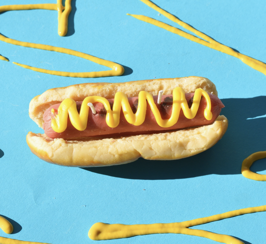 Candle shaped like a hotdog in a bun with yellow mustard squirted along the top. 