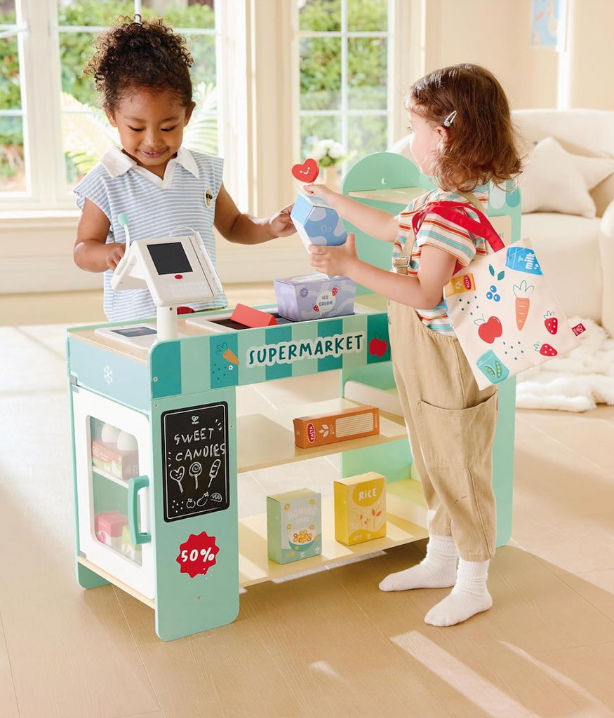 Two children playing at the wooden supermarket playset, one child is shopping while the other is ringing up the items. 