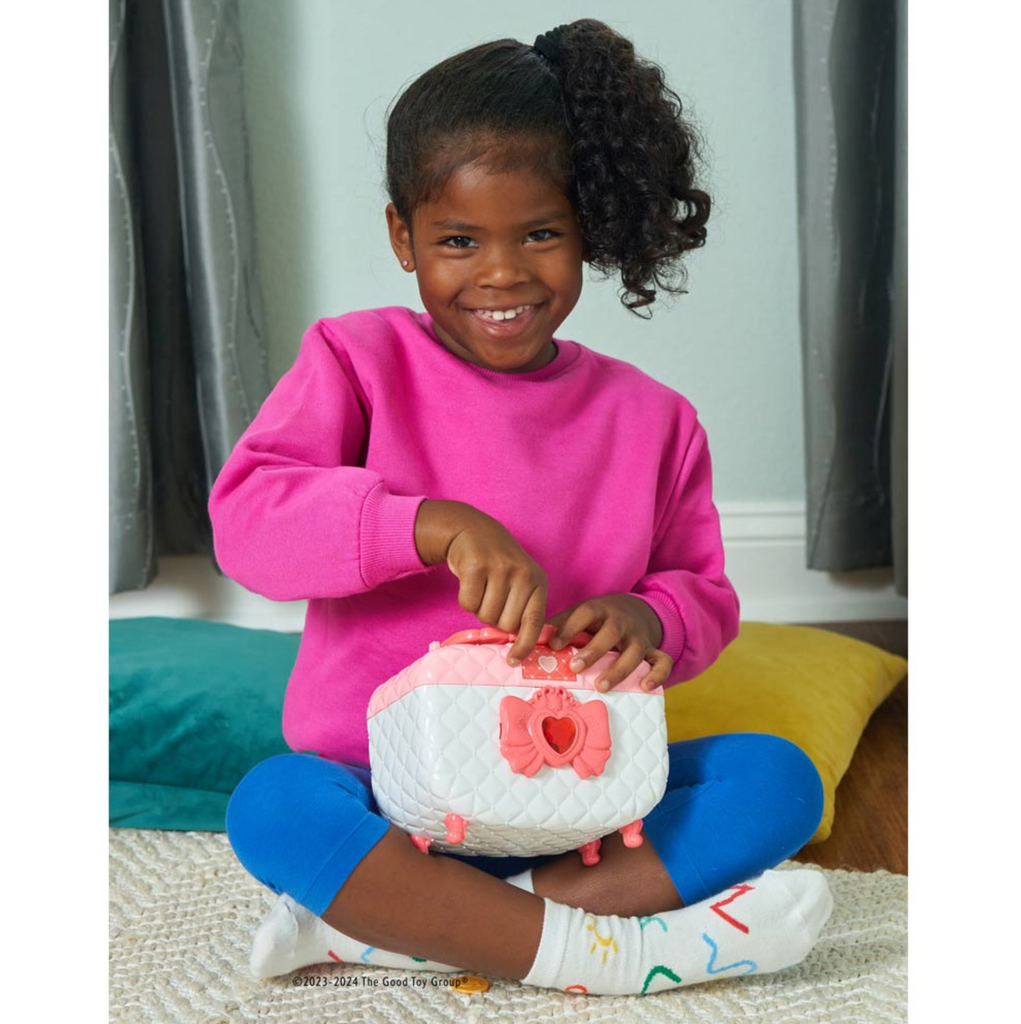 Smiling girl in a hot pink shirt and blue pants sitting on the floor holding the jewelry box safe.
